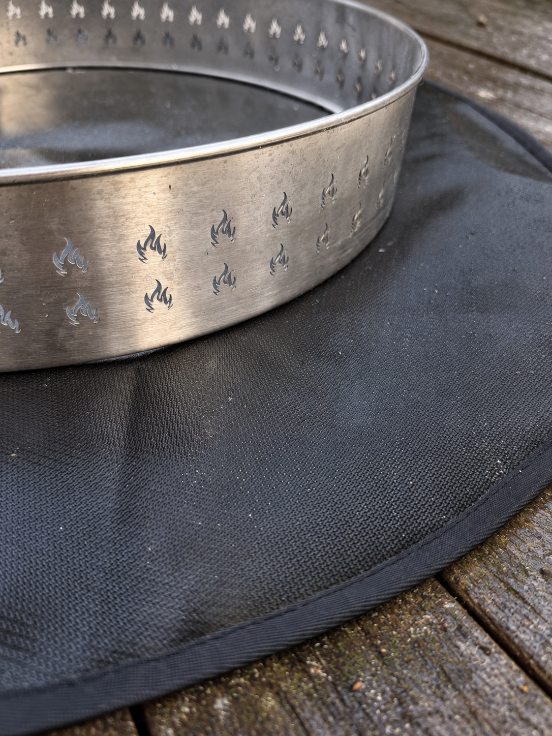 A silver metal fire ring with flame-shaped cutouts, resting on a black circular fire pad over a weathered wooden deck.