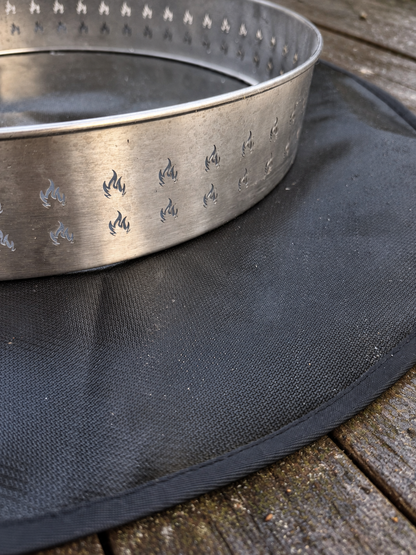 A silver metal fire ring with flame-shaped cutouts, resting on a black circular fire pad over a weathered wooden deck.
