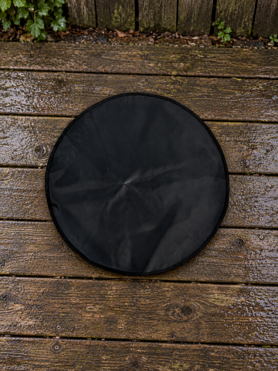 A round black heat mat cover with a black stitched edge, laid flat on a wet wooden deck outdoors.