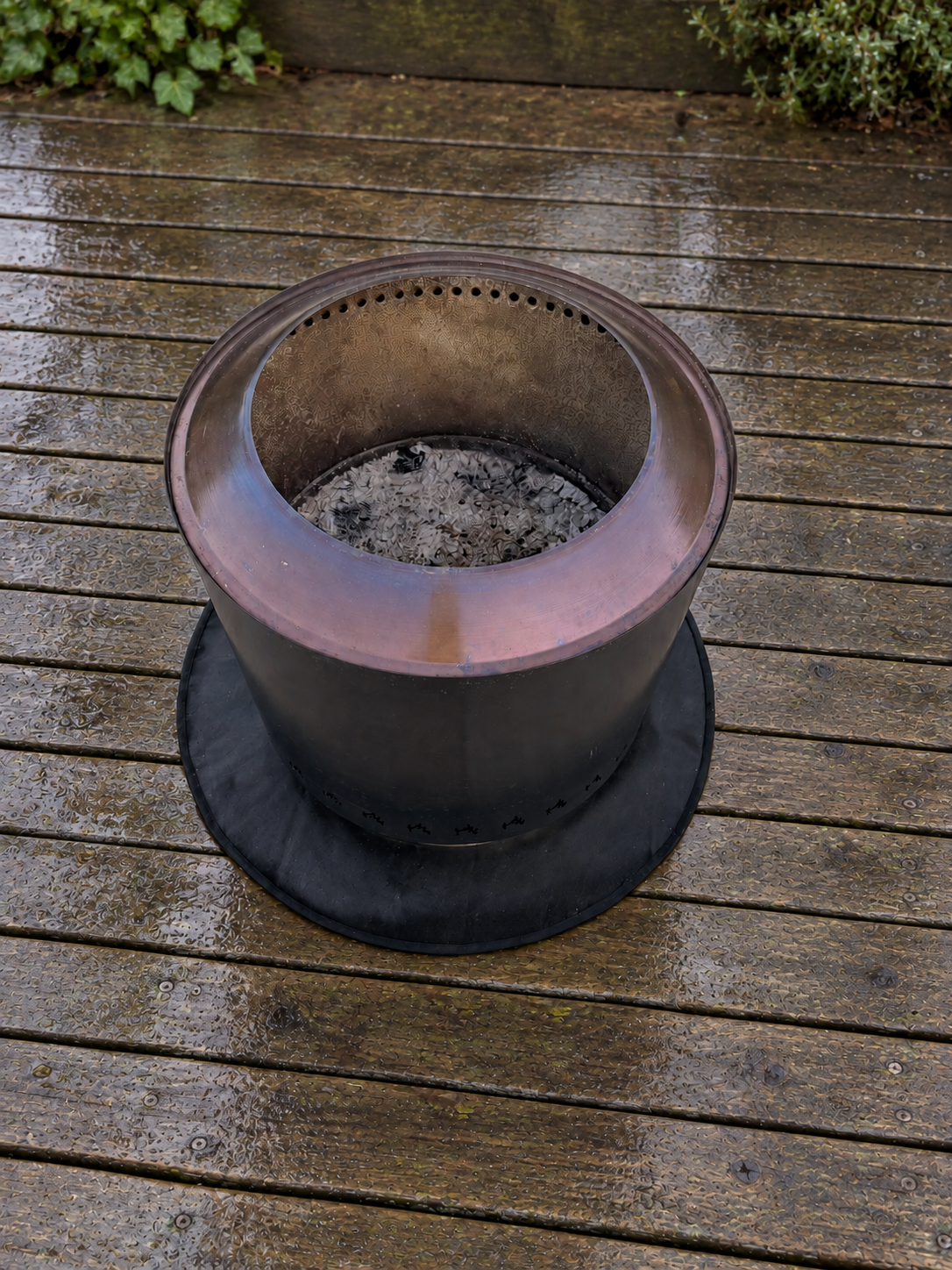 A stainless steel outdoor fire pit with a black heat mat base, shown on a wet wooden deck.